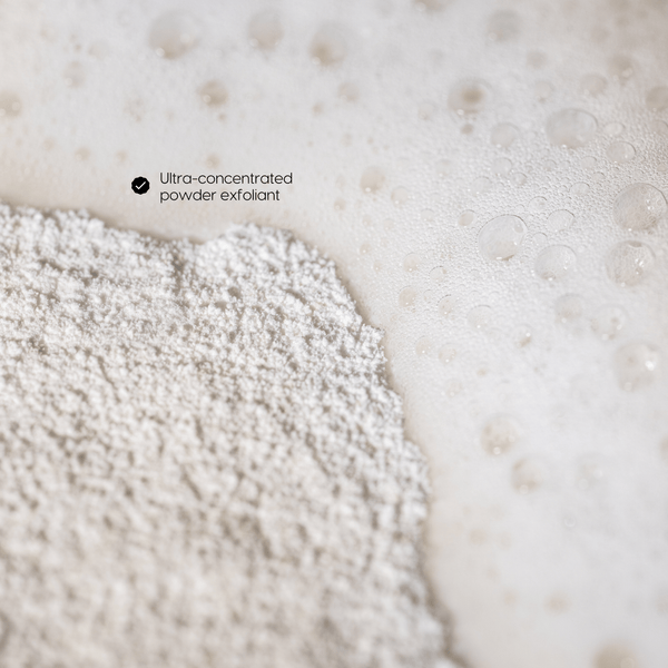 Close-up of exfoliating powder resting on a white foamy surface with small bubbles and the text 'Ultra-concentrated powder exfoliant' near the top left.