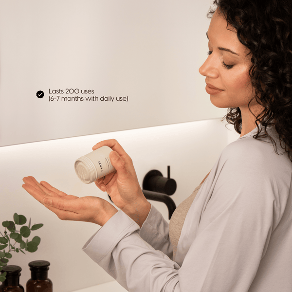 A woman in a bathroom holds a small skincare jar and pours the powder contents onto her hand. Text on the mirror reads 'Lasts 200 uses (6-7 months with daily use)'.