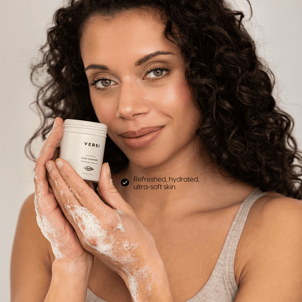 A woman with curly dark hair holds a beige cosmetic jar labeled Versi while applying a foaming cleanser to her hands. She is wearing a light beige tank top against a soft light gray background, with the text 'Refreshed, hydrated, ultra-soft skin.