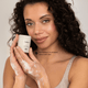 A woman with curly dark hair holds a beige cosmetic jar labeled Versi while applying a foaming cleanser to her hands. She is wearing a light beige tank top against a soft light gray background, with the text 'Refreshed, hydrated, ultra-soft skin.