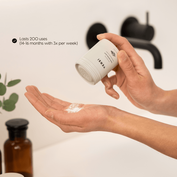 Person holding a beige container labeled 'Versi Face Exfoliant' over their palm with a blurred background.