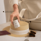 A hand holds a compact beige cosmetic jar labeled Verst over a beige travel bag, with a straw hat, sunglasses, and a passport nearby. Text on the image reads 'Compact & lightweight, perfect for travel'.