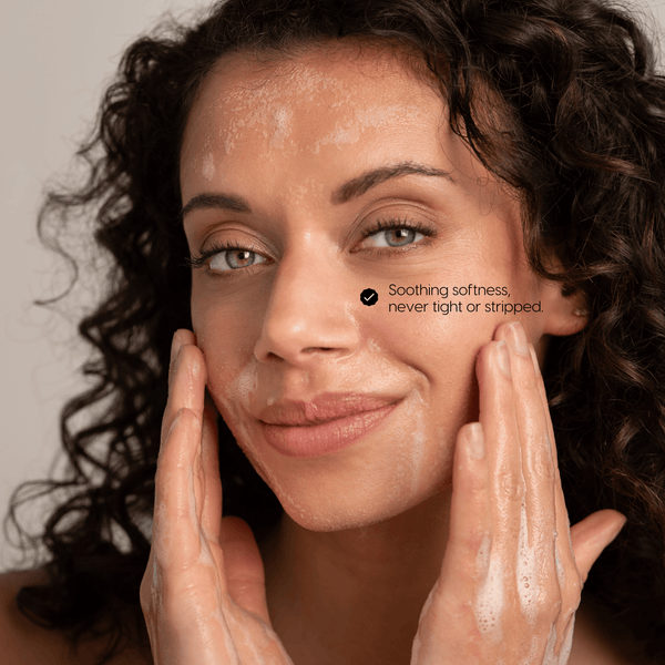 Close-up of a woman Chantal van Dalen with curly dark hair and bright eyes washing her face with foaming powder cleanser, touching her cheeks, with text reading 'Soothing softness, never tight or stripped.'.